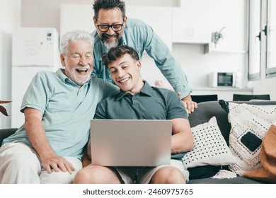 Happy multigenerational family of males watching funny videos on laptop together. Grandfather, mature son and young grandson are having fun and laughing while sitting on the sofa at home - Powered by Shutterstock