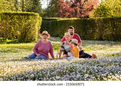 Happy Multigenerational Family In Straw Hats Having Fun During Picnic In Spring Blooming Garden On Sunny Day.