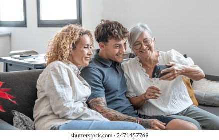 Happy multigenerational family watching funny videos on smartphone together. Grandmother, mature daughter and young grandson having fun and laughing while sitting on the sofa at home - Powered by Shutterstock