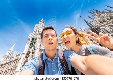 Happy Multinational Couple In Love Hugs And Takes A Selfie Photo On The Background Of The City Hall Tower In Munich. Honeymoon Trip To Germany