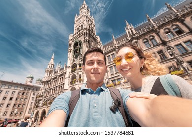 Happy Multinational Couple In Love Hugs And Takes A Selfie Photo On The Background Of The City Hall Tower In Munich. Honeymoon Trip To Germany