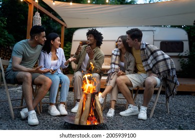 Happy multinational friends sitting near campfire, talking to each other, enjoying warm summer evening next to RV, spending time together on camping trip. Summertime tourism and recreation - Powered by Shutterstock