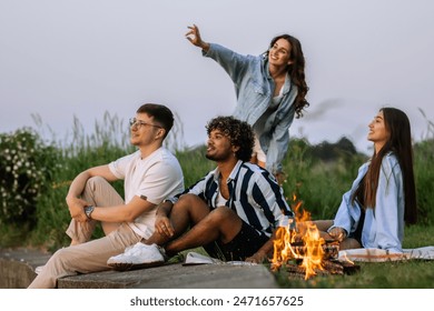 Happy multinational friends sitting near campfire, talking to each other, looking at the landscape and enjoying warm summer evening.  - Powered by Shutterstock