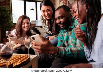 Happy Multiple Nationalities, Ethnicities Group Friends Looking, Watching Funny Messages At Smartphone Screen, Smiling, Laughing, Drinking Wine, Eating Bread Sticks At Apartment Reunion.