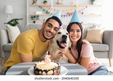 Happy multiracial couple celebrating their dog's birthday with festive cake, wearing party hats at home. Cute golden retriever having b-day with his owners, enjoying anniversary