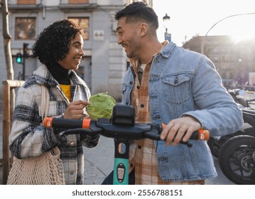 Happy multiracial couple is holding a lettuce and electric scooters while talking on a city street during a sunny day, celebrating valentine's day with a healthy lifestyle - Powered by Shutterstock