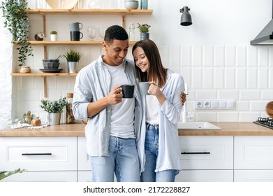 Happy multiracial couple in love, hispanic attractive man and pretty caucasian woman, stand at home in the kitchen, hug, drink tea or coffee together, enjoy spending time with each other, smile - Powered by Shutterstock