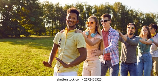 Happy Multiracial Friends Having Fun On Warm Sunny Evening In Park. Diverse Group Of Cheerful Joyful Young People Enjoying Outdoor Summer Party, Dancing In Conga Line All Together And Smiling