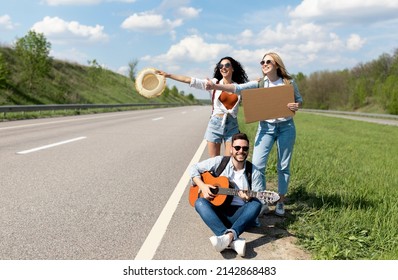 Happy multiracial friends hitchhiking, trying to stop car with empty carton sign, mockup. Group of millennial people with guitar waving down auto on highway. Traveling on summer vacation