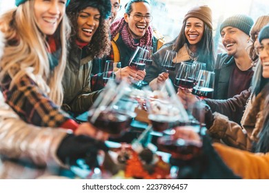 Happy multiracial friends toasting red wine at restaurant balcony - Group of young people wearing winter clothes having fun at winebar table - Dining life style and friendship concept