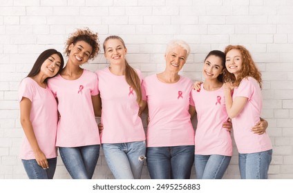 Happy Multiracial Ladies In Breast Cancer Pink T-Shirts With Awareness Ribbons Embracing Standing Near White Brick Wall Indoor - Powered by Shutterstock