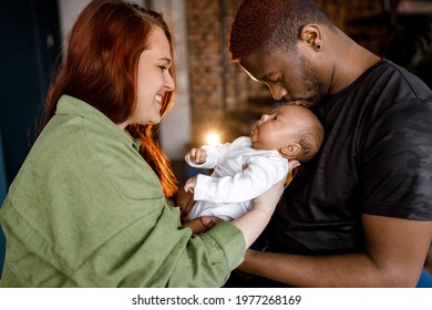 Happy Multiracial Mixed Family Lovely Holding Newborn Baby Son. Red Haired Caucasian Mom Looking At Her Child, Afro American Father Gently Kissing Cute Baby In Head. 