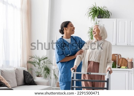 happy multiracial nurse in uniform laughing with retired woman while standing near walking frame at home