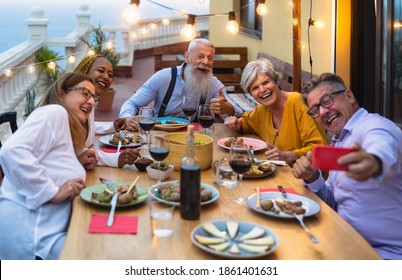Happy multiracial senior friends having fun dining together while taking selfie with mobile smartphone on house patio - Elderly lifestyle people and food concept 