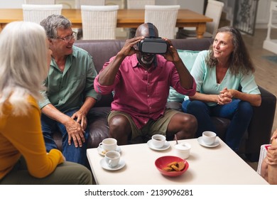 Happy Multiracial Senior Male And Females Looking At Friend Using Vr Headset At Home. Unaltered, Lifestyle, Social Gathering, Friendship, Virtual Reality, Futuristic And Modern Technology.