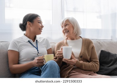 happy multiracial social worker having tea and chatting with senior woman in living room - Powered by Shutterstock