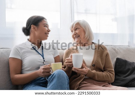 happy multiracial social worker having tea and chatting with senior woman in living room
