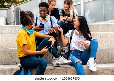 Happy Multiracial Teenage High School Students Sit On Steps Talking And Using Mobile Phones Outside.