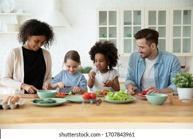 Happy Multiracial Young Family With Little Daughters Cooking Healthy Salad In Kitchen Together, Smiling Caring Diverse Parents Have Fun Enjoy Weekend With Small Kids, Prepare Food Or Breakfast At Home