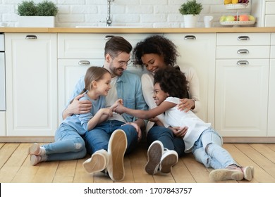 Happy multiracial young parents relax on warm wooden floor in kitchen with little daughters, smiling family with small girls children sit rest in modern design renovated home, have fun together - Powered by Shutterstock