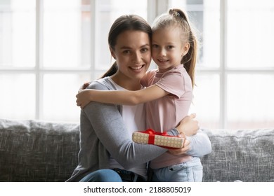 Happy Mum And Cute Preschooler Daughter Girl Celebrating Mothers Day At Home. Millennial Mommy Receiving Birthday Gift Wrap From Kid, Hugging Child, Looking At Camera And Smiling. Family Concept