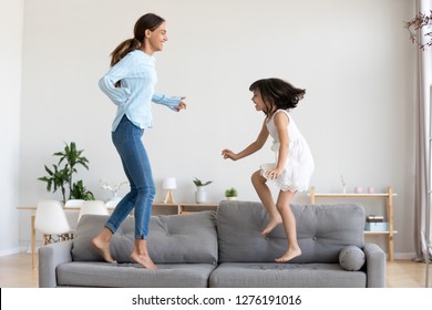 Happy Mum And Little Daughter Playing Jumping On Comfortable Sofa In Living Room, Mother Baby Sitter Laughing Having Fun With Cute Kid Girl At Home, Happy Mom And Child Enjoy Funny Activity Together