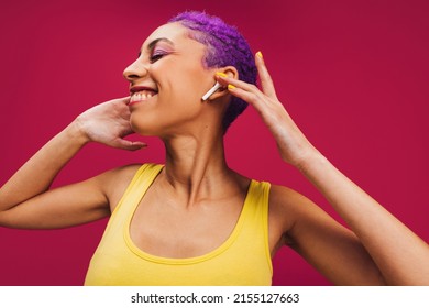 Happy music only. Carefree young woman touching her wireless earphones while listening to her favourite song. Woman with purple hair smiling cheerfully against a pink background,