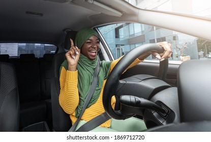 Happy muslim black female driver in hijab singing and dancing while driving car in city, cheerful islamic lady in headscarf having fun and enjoying road trip with her new vehicle, free space