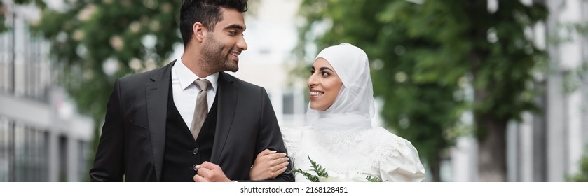 happy muslim bride in wedding hijab and white dress holding bouquet near groom outside, banner