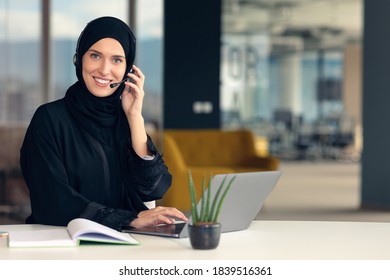 Happy muslim businesswoman in hijab at office workplace. Smiling Arabic woman working on laptop and talking on smartphone