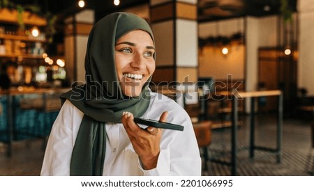 Happy Muslim businesswoman smiling while speaking on the phone in a cafe. Cheerful woman with a hijab communicating with her clients while working remotely.