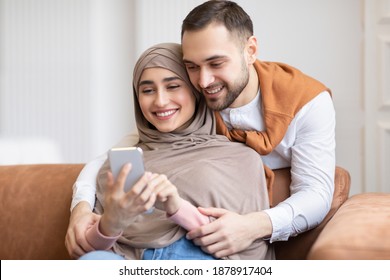 Happy Muslim Couple Using Smartphone Texting And Browsing Internet Sitting On Couch At Home. Family Leisure. Cheerful Arab Spouses Watching Video Online On Phone. Great Mobile Application Concept