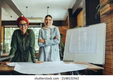 Happy Muslim designers smiling at the camera while working on blueprint drawings. Two creative businesswomen planning a new project in an office. Architects wearing headscarfs in a modern workplace.