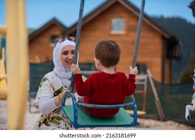 A Happy Muslim Family Is Having Fun In The Park Arab Mother In Hijab Pushes Child Son On A Swing In The Garden Playground.	