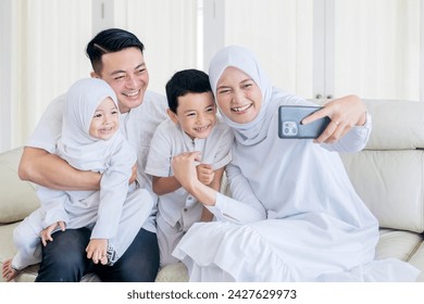 Happy muslim family making a video call or taking a selfie while sitting together on the couch during Eid Mubarak in the living room - Powered by Shutterstock