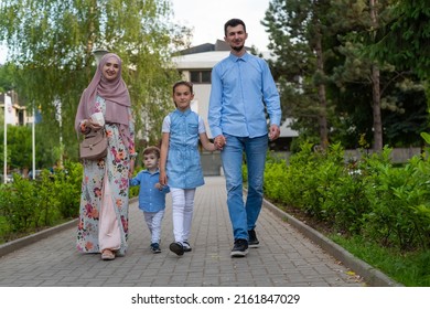 Happy Muslim Family Mother And Father Walking And Holding Hands With Two Children In The Summer Park.