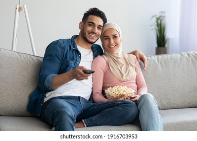 Happy muslim family of two watching TV together at home, hugging and eating popcorn, copy space. Joyful middle-eastern man and woman having fun together on weekend, love and relationships concept