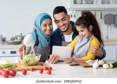 Happy Muslim Family Using Digital Tablet While Cooking In Kitchen Together, Cute Little Arab Girl And Her Islamic Parents Checking Online Recipe On Tab Computer While Preparing Food At Home