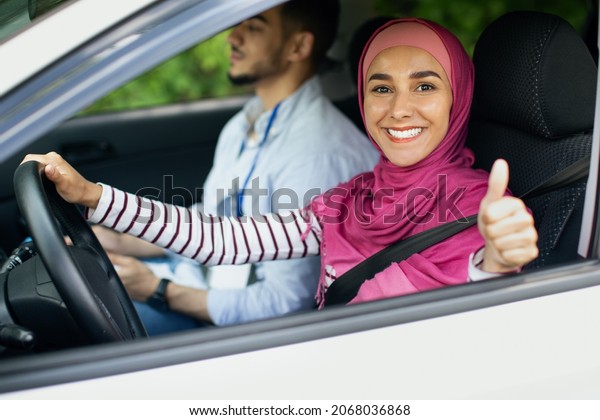 Happy Muslim Female Driver Showing Thumb Up After\
Successful Exam Passing In Driving School, Cheerful Middle Eastern\
Woman In Hijab Sitting In Car Next To Instructor And Smiling At\
Camera, Closeup