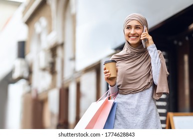 Happy muslim girl with shopping bags drinking coffee and talking on phone, walking by street, empty space