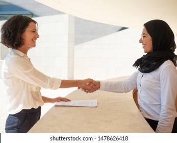Happy Muslim hostess greeting cheerful female tourist in hotel. Caucasian and Arab women in formal suits shaking hands over reception desk. Reception desk concept