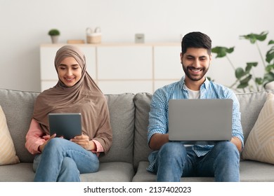 Happy Muslim Husband And Wife Browsing Internet On Laptop And Digital Tablet Sitting On Sofa At Home. Middle Eastern Family Couple Using Gadgets Spending Weekend Indoors. Technology Lifestyle