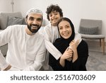 Happy Muslim kid boy and cheerful parents in Arab clothes posing for portrait on home floor, hugging, laughing, looking at camera with toothy smiles. Mom on hijab piggybacking kid, having fun