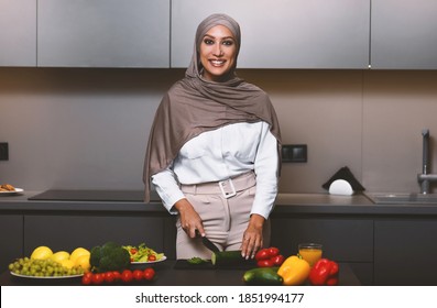 Happy Muslim Lady Cooking In Kitchen, Cutting Fresh Vegetables Making Salad For Dinner At Home, Wearing Hijab. Arab Wife Preparing Healthy Meal Smiling To Camera Indoors. Arabic Cuisine And Recipes