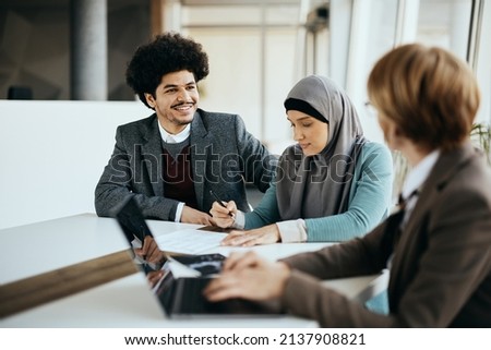Happy Muslim man talking to financial advisor while his wife is signing paperwork during the meeting in the office. 