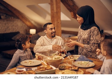 Happy Muslim Man Talking To His Wife Who Is Pouring Water Into His Glass During Family Meal At Dining Table. 