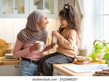 Happy Muslim Mom In Hijab And Her Little Daughter Bonding Together In Kitchen, Islamic Family Talking And Laughing, Mother Enjoying Coffee While Her Female Child Sitting At Table And Drinking Milk