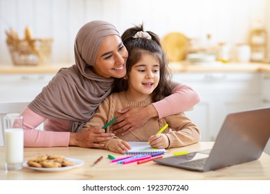 Happy Muslim Mom In Hijab And Her Little Daughter Using Laptop In Kitchen, Having Fun Together At Home, Watching Cartoons Or Kids Development Videos Online, Small Girl Is Drawing At Table, Free Space - Powered by Shutterstock