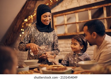 Happy Muslim Mother Pouring Water Into Daughter's Glass During Family Meal At Home.