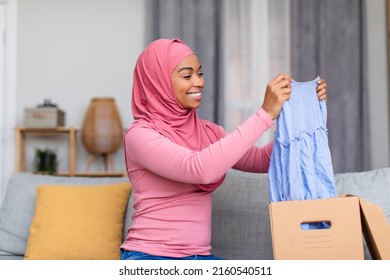 Happy muslim shopaholic lady receiving new outfit from shop, holding new clothes, unpacking cardboard box, excited about successful shopping, sitting on sofa at home